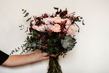Una mano sujetando un ramo de flores en tonos rosas y granates. Eucalipto plateado, crisantemo,...