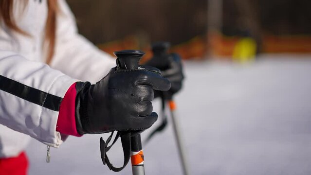 Close-up ski poles handle with female hands in leather gloves taking equipment leaving in slow motion. Unrecognizable fit young woman walking away on sunny day on winter resort