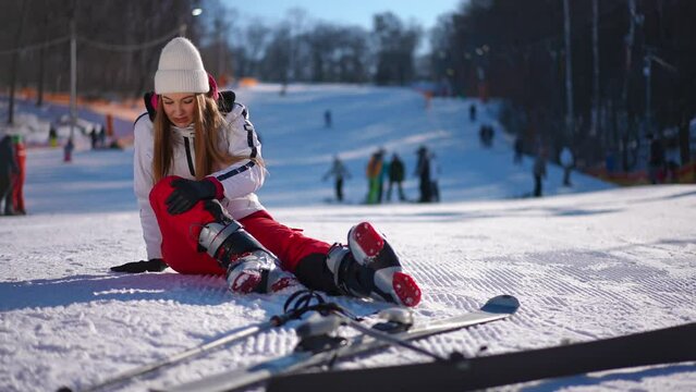 Beautiful fit sportswoman sitting on white snow caressing painful knee. Wide shot portrait of sad fallen Caucasian woman on winter resort on sunny day