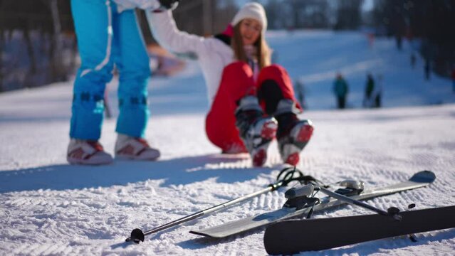 Skis on white snow with blurred woman with pain in knee standing up with help of friend at background. Young slim Caucasian lady having injury skiing on winter resort on sunny day