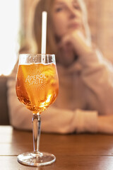 A glass of Aperol and a slice of orange on a table in a cafe. In the background, a blurry figure of a woman sitting at a table