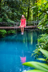 Fototapeta na wymiar a girl in a red dress sits on a bridge over the blue rio celeste river in volcano tenorio national park; sky blue river surrounded by dense rainforest in costa rica