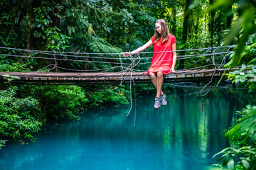 Fototapeta na wymiar a girl in a red dress sits on a bridge over the blue rio celeste river in volcano tenorio national park; sky blue river surrounded by dense rainforest in costa rica