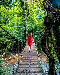 girl in red dress walks across suspension bridge in dense jungle in volcano tenorio park by rio celeste river; walking through rainforest in Costa Rica