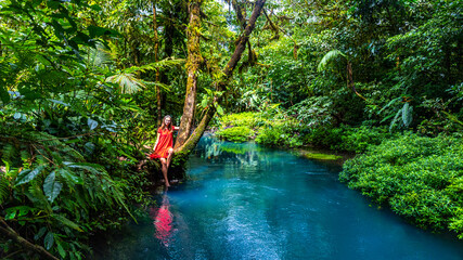 girl in red dress stands on the banks of the famous blue rio celeste river in volcano tenorio national park  unique blue river surrounded by dense tropical rainforest © Lens Down Under