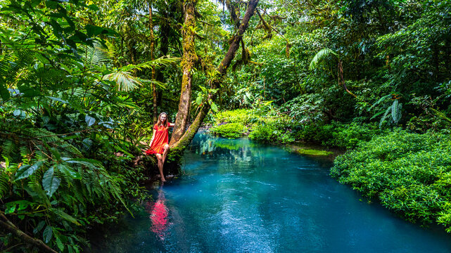 girl in red dress stands on the banks of the famous blue rio celeste river in volcano tenorio national park; unique blue river surrounded by dense tropical rainforest