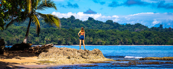 A beautiful girl stands on the rocks under a palm tree on the shore of a Costa Rican beach in the...