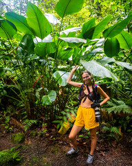 girl photographer stands under big green leaves in Costa Rican tropical rainforest; hiking through the jungle in Costa Rica