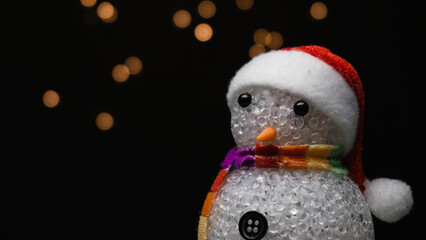 Figurine of snowman with Santa Claus hat on black background with orange bokeh lights
