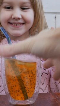 caucasian cute preschool girl got a full glass of mango juice with passionfruit seeds and ice cubes. A girl drinks a natural fruit drink through a reusable straw. Vertical video, dolly shot