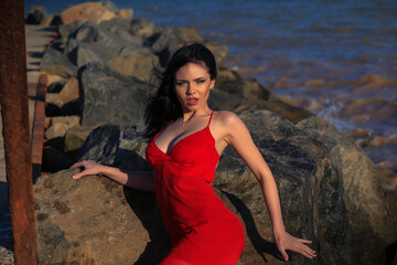 a beautiful nice young fashionable girl with black hair in a red dress stands in summer by the water of the sea ocean and stones