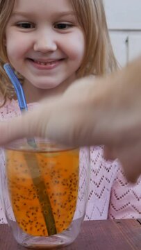 a little Caucasian girl tastes mango juice with chia seeds and ice cubes for the first time and is surprised by the taste. The child drinks a drink through a reusable drinking tube. slow motion