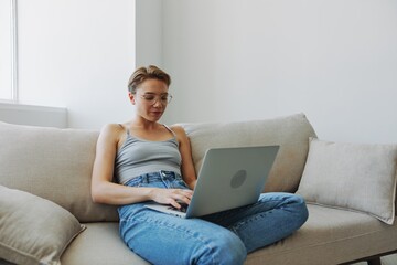 Teenage girl freelancer with laptop sitting on couch at home smiling in home clothes and glasses with short haircut, lifestyle with no filters, free copy space