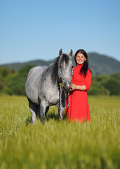 Woman with dark hair and bright red dress walking her Arabian gray horse in green field on sunny day, smiling as animal eats grass