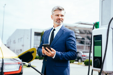 Handsome mature grey haired man in formal wear holding smartphone while charging car at electric vehicle charging station outdoor.