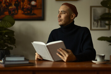 A caucasian man wearing a beanie and a knitted sweater sitting at a wooden table in a living room reading a book. 