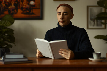 A caucasian man wearing a beanie and a knitted sweater sitting at a wooden table in a living room reading a book. 