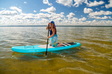 A Jewish woman in a kerchief kisuy rosh on her head and in a pareo on her knees on a SUP board with an oar swims in the lake on a sunny day.