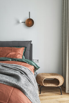 Bedroom interior with rust color linen and cushions on a bed, grey blanket, rattan bedside table and metal wall light.