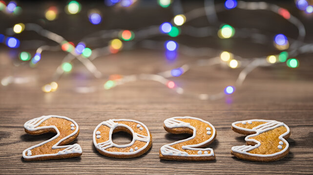 Beautiful New Year card for happy 2023 from ornate gingerbread numerals and LED light strings. Closeup of sweet cookies of Xmas pastry with blurry colored electric bulbs on wood background with bokeh.