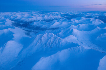 Aerial view of the Yukon during the winter with a pink glow in the sky at sunrise over the snow covered mountain peaks creating a surreal, meringue-like landscape below; Yukon, Canada