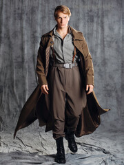 hero in a military style with a gun in his hands, a young man in a long brown coat and breeches with suspenders. Photo in the studio on a gray background - 552685546