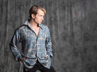 Stylish elegant young man in a blue silk shirt posing in the studio on a gray fabric background