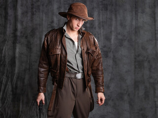 A young man in retro style, an adventure character. The hero of the adventure, a guy in a hat and a leather jacket, posing in the studio on a gray background