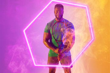 Smiling african american male tennis player holding racket amidst smoke over illuminated hexagon