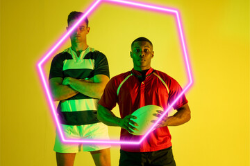 Multiracial confident male rugby opponents with arms crossed and ball standing by glowing hexagon