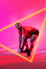 African american male player with rugby ball bending by illuminated triangle on pink background
