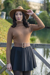 Thoughtful woman sitting on the stairs in a brown hat and autumn foliage on the river bank.