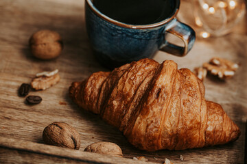 Ceramic cup of black coffee and croissant on wooden tray with walnuts, coffee beans and lights close up