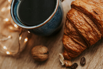 Porcelain mug of black coffee with walnuts and croissant on wooden tray - background with lights