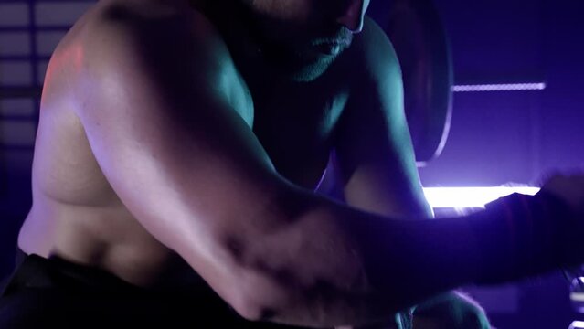 Muscular male bodybuilder with a naked torso wrapping bandages for boxing around his hands. Man sitting in gym against background of rack with dumbbells and barbells. Close up in blue light.