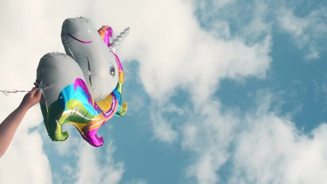 Vertical video of hand holding a colorful unicorn balloon on cloudy sky at background