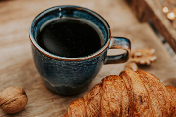 Cup of black hot coffee on wooden tray with walnuts and delicious croissant closeup