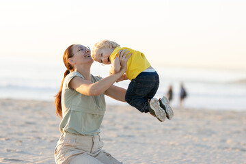 Fototapeta na wymiar Smiling ginger woman raising up her son sitting on the beach