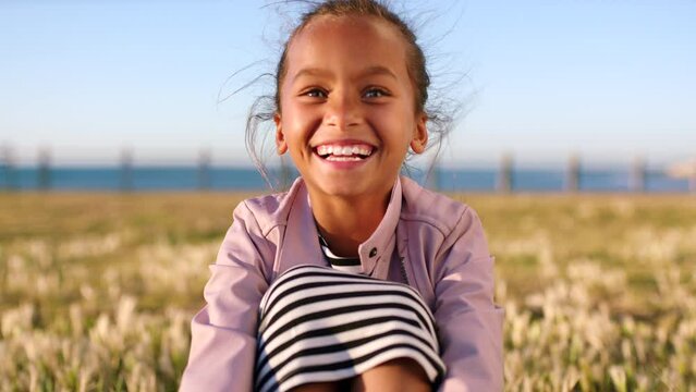 Girl, kid and laughing in park, promenade and nature in summer, sunshine and outdoor environment in Colombia. Portrait of happy, smile and funny child relax on grass lawn by sea wind for happiness