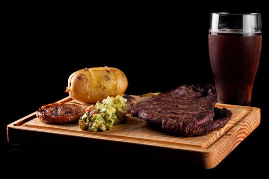 Corte de arrachera con chistorra, papa al horno, servido en una tabla, acompa&ntilde;ado de cerveza fr&iacute;a oscura en una copa.