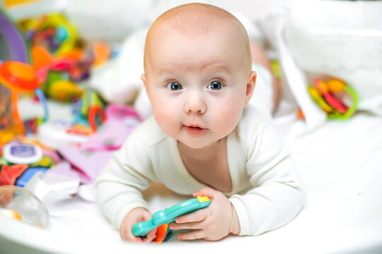 A funny baby in white clothes lies on a cot near toys. A cute little newborn girl lies on a bed among colorful toys for babies. The first educational toys for sensory development.