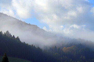 Niedrige Wolke schmiegt sich an einen bewaldeten Berg. Der Hintergrund ist neblig, der Himmel blau. 