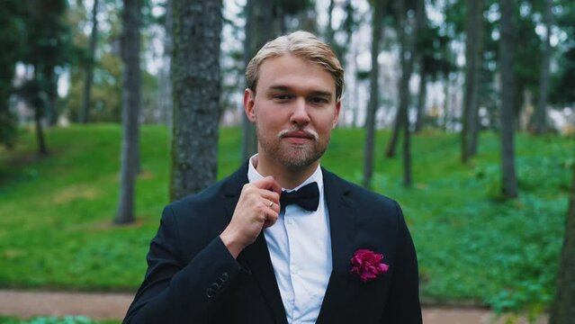 Good-looking fit blond bearded Scandinavian groom in his late 20s correcting his black butterfly tie and looking at camera. Blurred forest in the background. High quality 4k footage