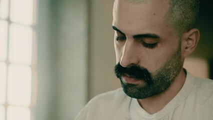 Close-up face of a thoughtful, serious, and focused handsome bearded Caucasian young male businessman working at home office, writing plans, thinking, and brainstorming ideas. Background window.