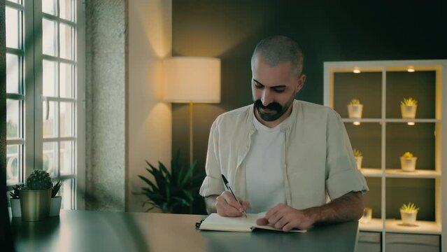 Good-looking motivated Caucasian biographer or novelist author handwrites a biography, romance, or novel in a small notebook. Inspired, attractive screenwriter uses a journal to write a screenplay.