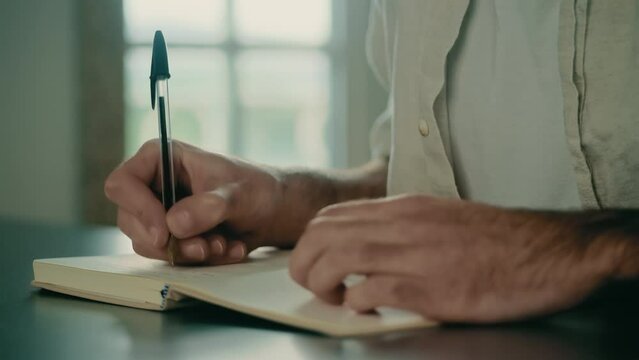 Male hands holding a pen, writing, and journaling in a small notebook. Writer, author, biographer, dramatist, journalist, novelist, screenwriter, or columnist writing in a journal. Close-up view.