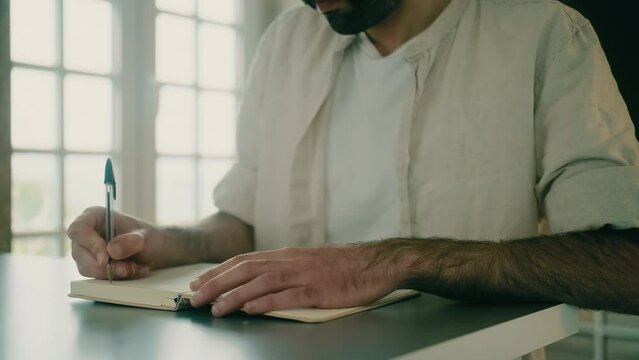 Close-up view of a male creative freelancer sitting at a desk, holding a pen, and taking notes. A writer sitting at a table finishes his work, closes his notebook, stands up, and leaves the room.