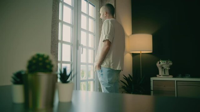 A frustrated Caucasian young man with a mustache wearing jeans and a cream shirt stands at a window in the apartment's living room and looks outside while thinking about what he should do next.