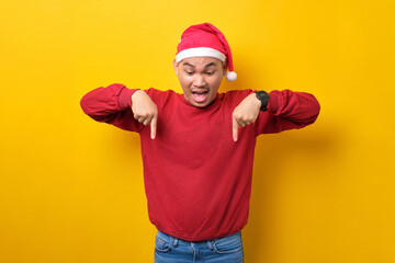 Naklejka premium Surprised young Asian man in Santa hat pointing fingers down, demonstrating free copy space for advertisement on yellow studio background. celebration Christmas holiday and New Year concept