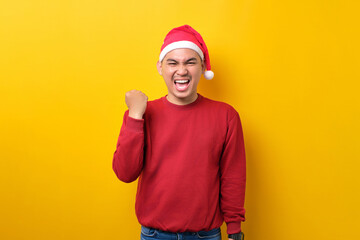 Excited young Asian man in Santa hat raising fists up, celebrating success over yellow studio...